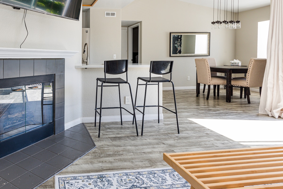 A spacious open-plan living area is highlighted by a cozy fireplace and modern bar stools positioned at the counter. The dining area features a dark wood table surrounded by upholstered chairs, while natural light floods the room through large windows, illuminating the stylish floor.