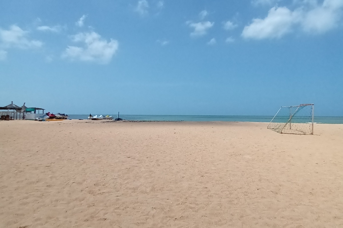 A wide expanse of sandy beach is shown, with clear blue skies and calm ocean waters in the background. A goalpost is positioned on the right side of the image, while colorful boats are docked in the distance. Light clouds are visible above.
