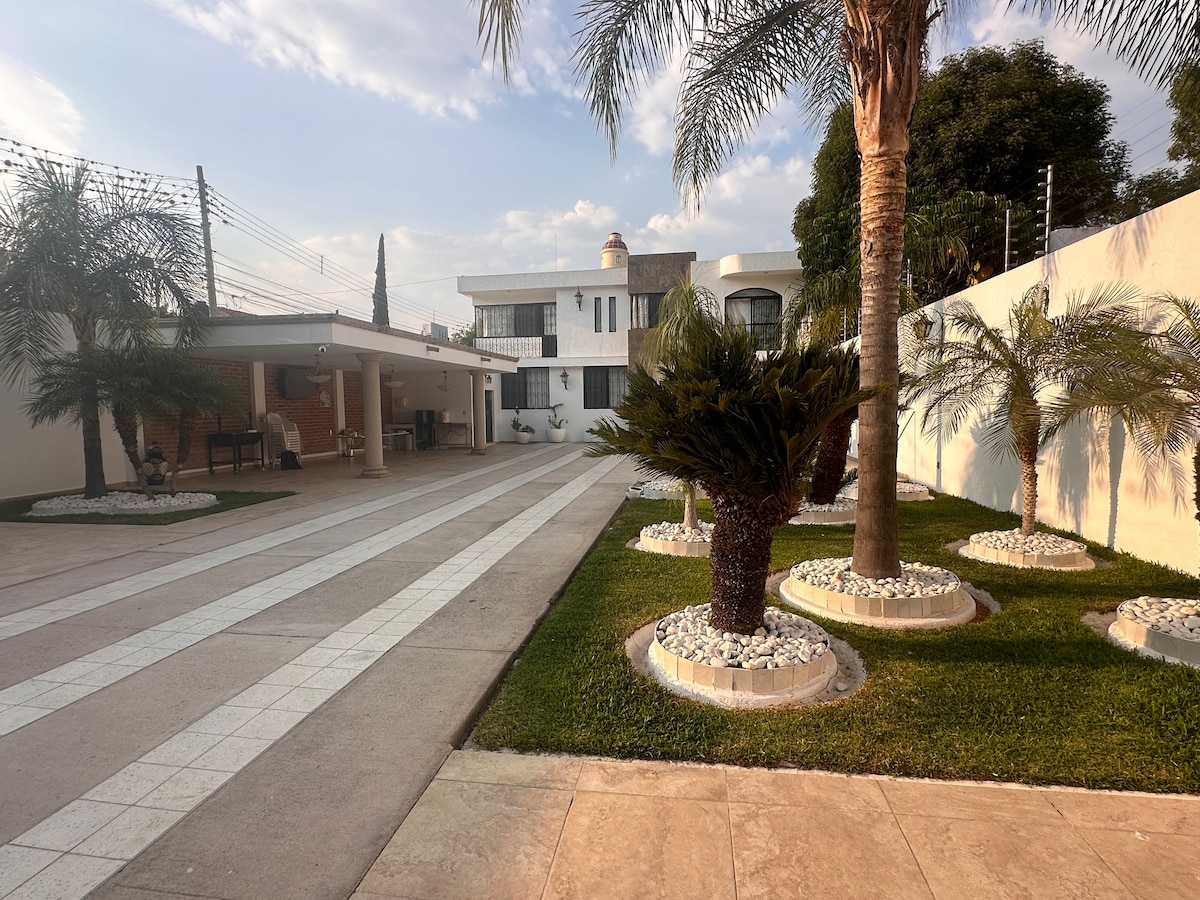 A spacious outdoor area features a well-maintained driveway lined with white stone pathways and lush green grass. Palm trees are planted in decorative beds, creating a welcoming entrance. The modern architectural style of the house is complemented by its surrounding tranquil setting.
