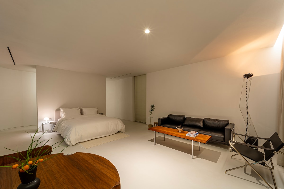 A spacious room features a soft, neutral color palette, with a large bed dressed in white linens. A sleek black sofa sits across from a modern wooden coffee table, complementing minimalistic decor. Soft lighting from the ceiling creates a calm ambiance.