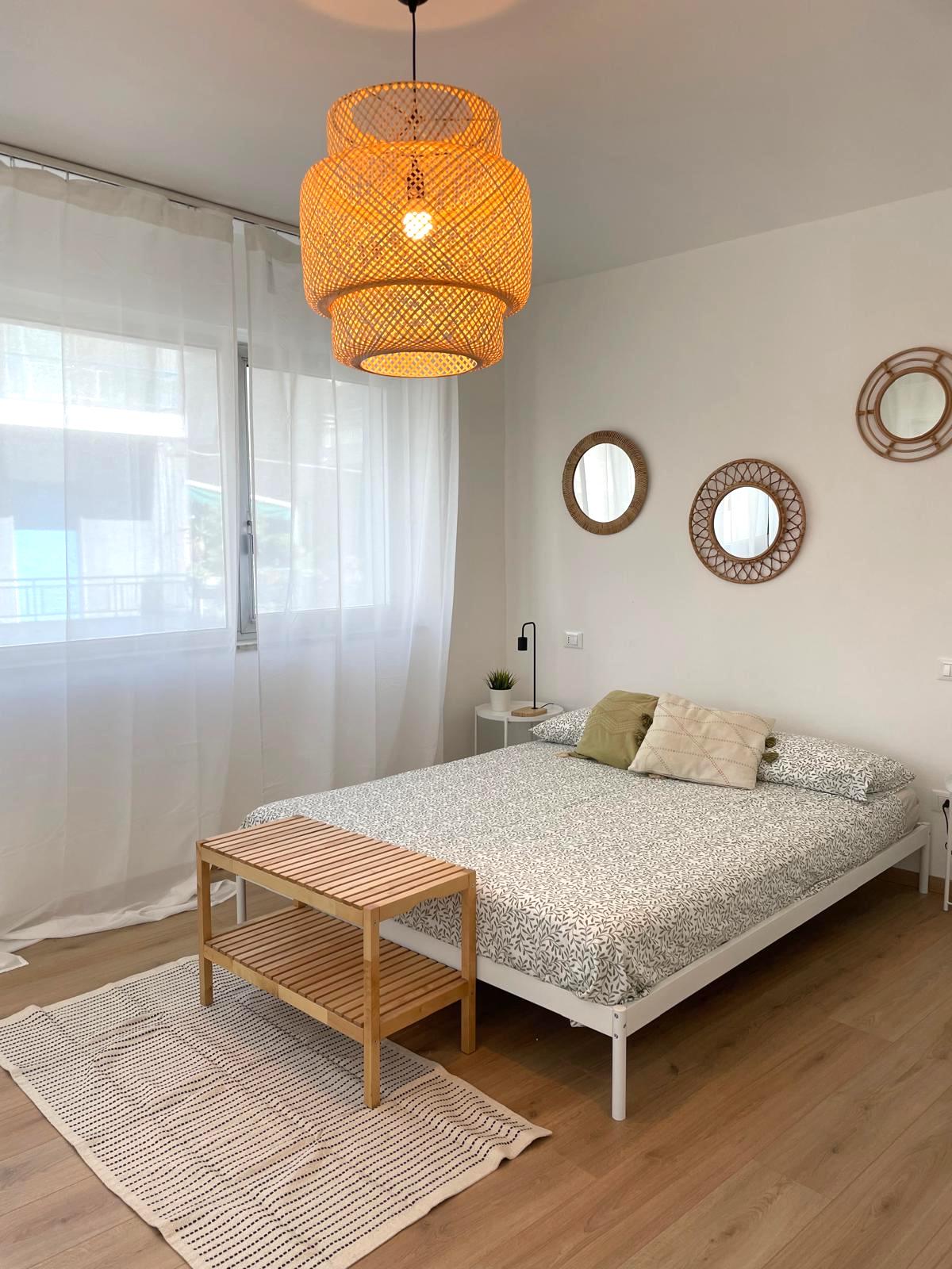 A bright bedroom features natural light streaming through large windows, with sheer curtains offering a soft drape. A comfortable bed is adorned with a patterned duvet and decorative pillows, complemented by a simple wooden bench and a striped rug on the floor. Three round mirrors hang on the wall.