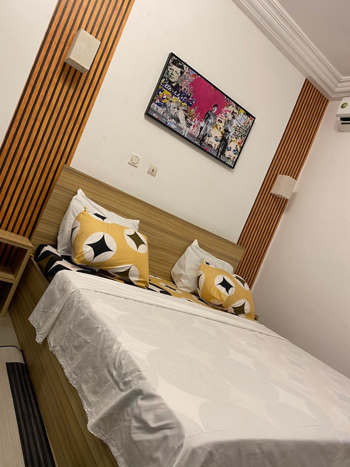 A cozy sleeping area features a spacious bed adorned with patterned throw pillows. The walls are accented with warm wooden panels and a vibrant piece of art. Wall sconces provide subtle lighting in the room, creating a functional and inviting space.