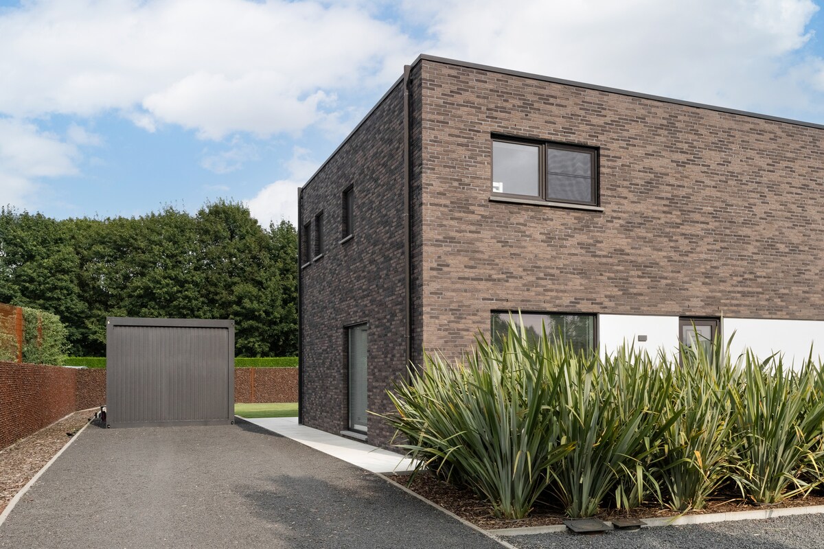A modern half-open home is displayed, featuring a dark brick exterior. A covered parking space is visible next to the house, while lush greenery surrounds the front. A clear blue sky and scattered clouds add to the serene setting.
