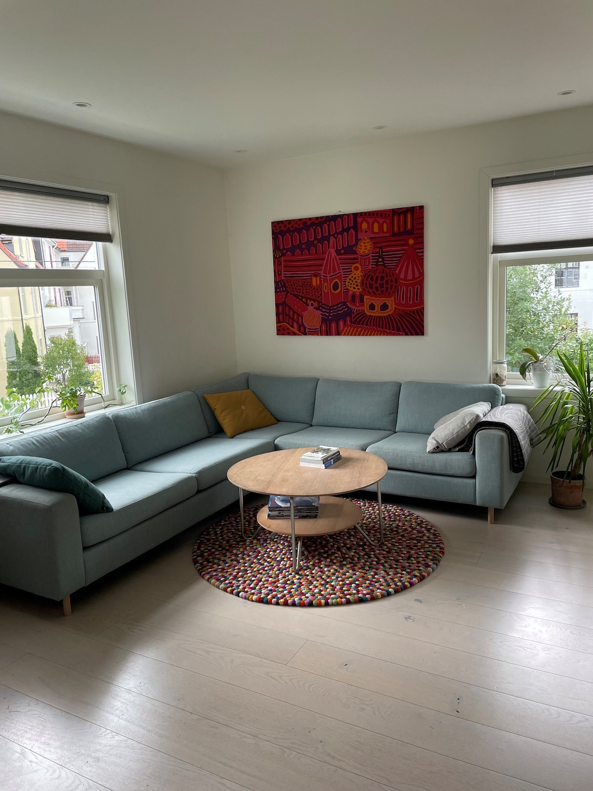 A bright living area is presented, featuring a light-colored sectional sofa adorned with decorative cushions. A round coffee table sits at the center, surrounded by a colorful rug. Windows provide natural light, and a potted plant adds a touch of greenery to the space.