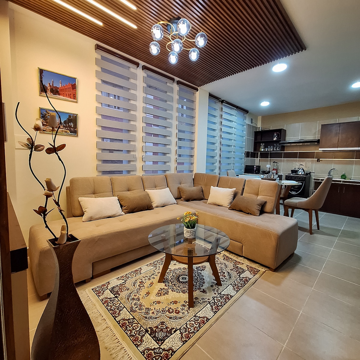 A spacious living area showcases a modern brown sectional sofa adorned with light cushions, set around a circular glass coffee table. A decorative vase rests on the table, and light walls are complemented by patterned window shades. The open kitchen area is visible in the background.