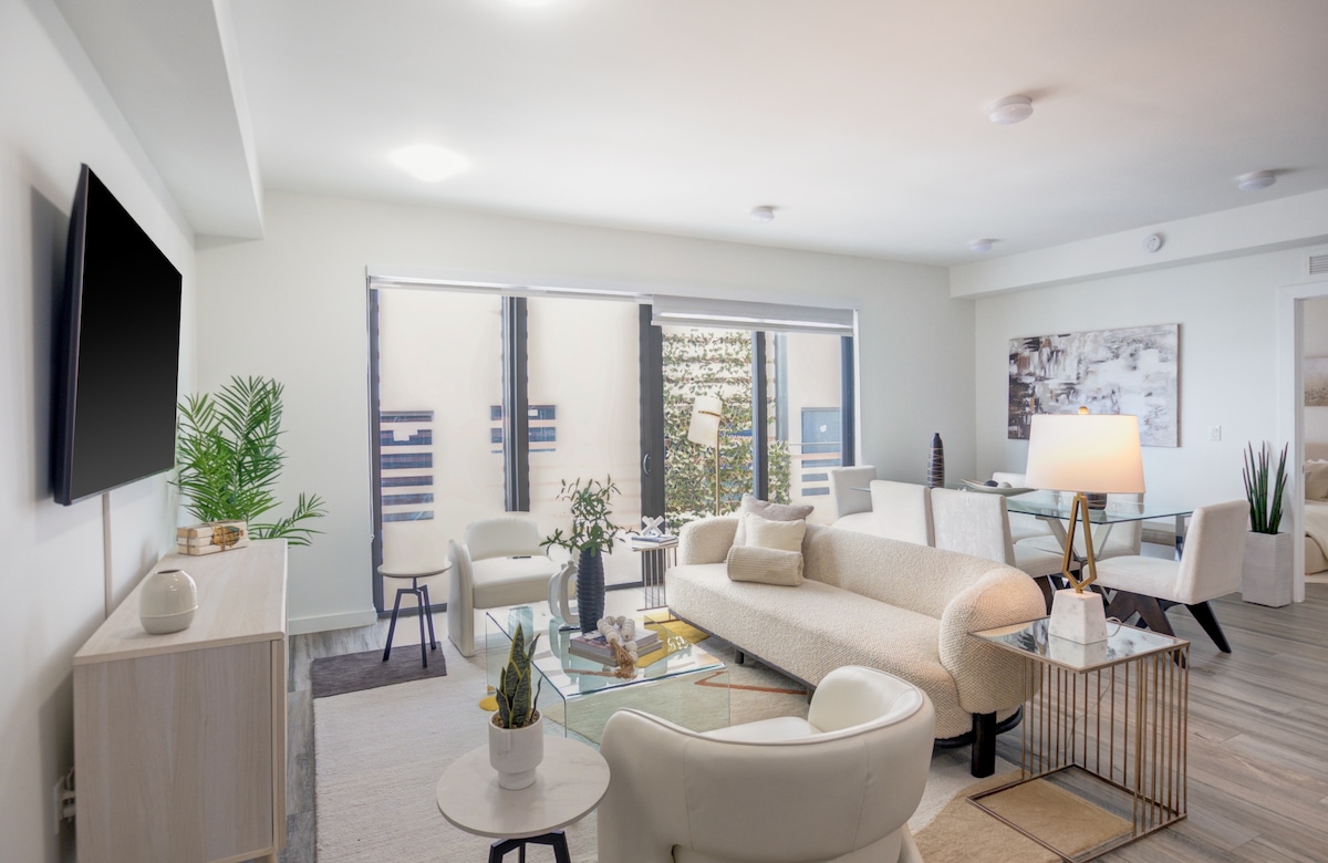 An open-concept living area features a modern design with a light-colored sectional sofa and glass coffee table. Natural light floods in through large sliding doors, leading to a private outdoor space. Decorative plants add freshness to the inviting atmosphere.