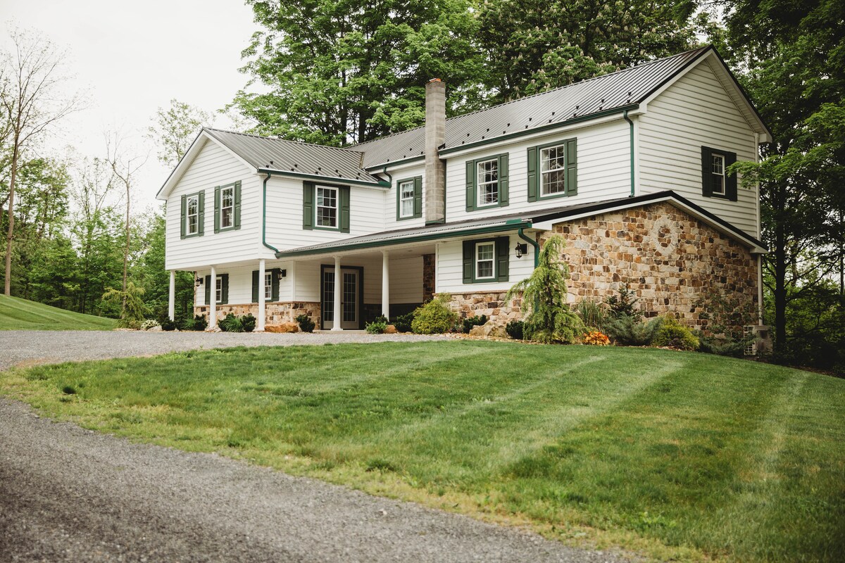 The exterior of a two-story farmhouse showcases a combination of white siding and stone. Green shutters frame the windows, and a wide pathway leads to the front entrance. Lush green grass surrounds the home, highlighting its natural setting among the trees.
