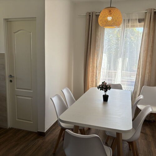 A dining area features a large white table surrounded by six sleek white chairs. A small potted plant is centered on the table. Light enters through a window with sheer curtains, and a woven pendant light hangs overhead, enhancing the bright and airy atmosphere.