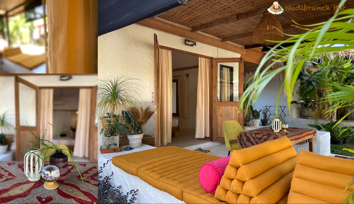 A cozy outdoor living space features a soft, yellow seating area with cushions arranged for relaxation. Potted plants add greenery, while a small table displays decorative items. Open doors invite natural light, enhancing the seamless connection between indoors and outdoors.