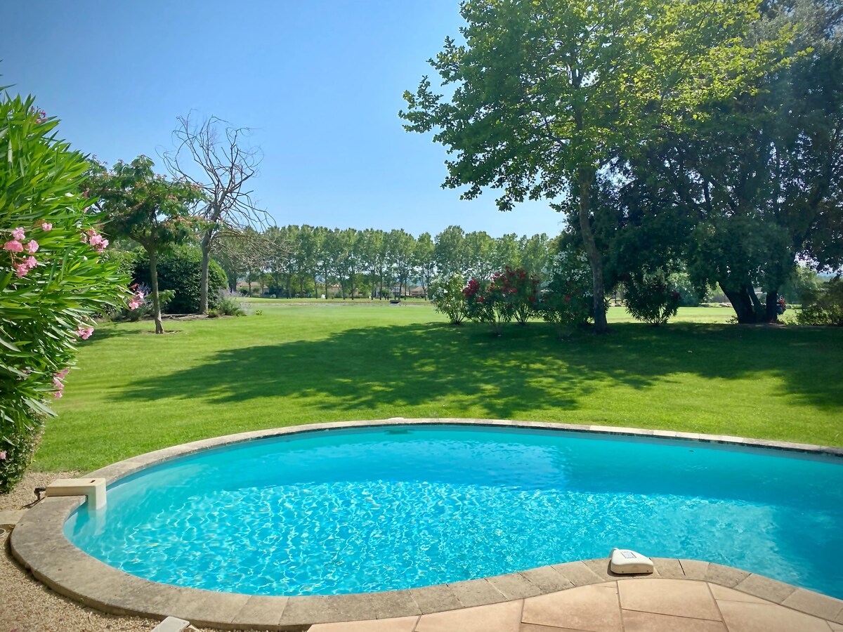 A private swimming pool is set amidst a well-manicured garden, featuring vibrant greenery and flowering plants. The pool's clear blue water contrasts with the serene landscape, which includes neatly trimmed grass and distant trees lining the perimeter.
