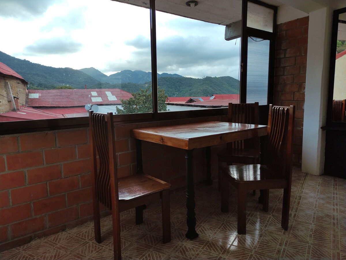 A wooden dining table with two matching chairs is positioned near large windows, providing views of the surrounding mountains and rooftops. Natural light illuminates the area, highlighting the brick walls and tiled floor.
