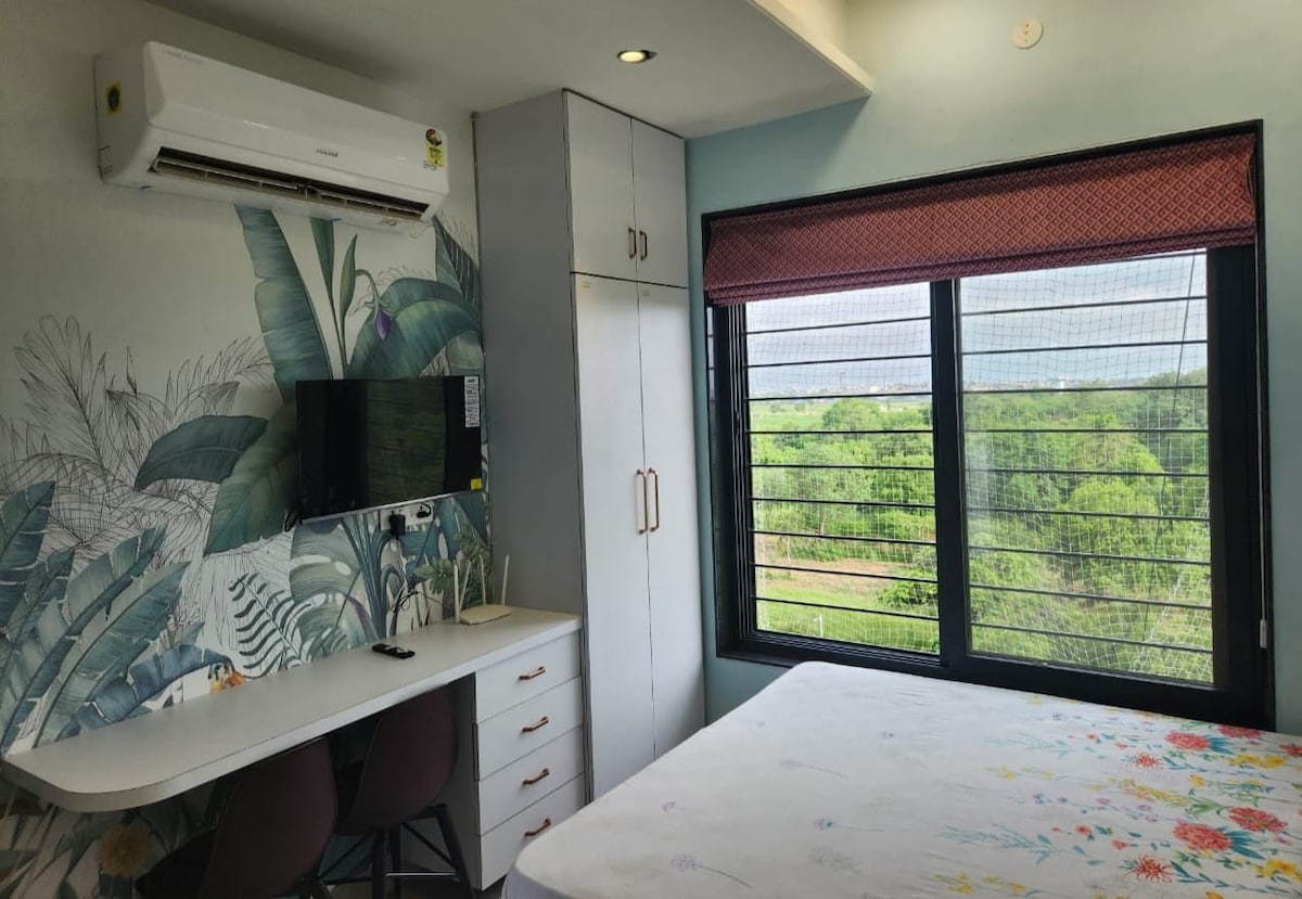 A bright bedroom is decorated with floral wallpaper and features a spacious bed. A desk with two chairs is positioned near a large window, which offers views of greenery outside. An air conditioning unit is mounted above the bed for comfort.