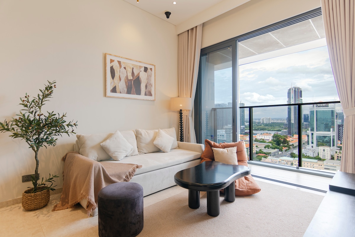 A modern living room is presented, featuring a light-colored sofa adorned with decorative pillows. A round black table sits in front of the sofa, complemented by a small stool. Floor-to-ceiling windows showcase a panoramic city view, enhancing the inviting ambiance.