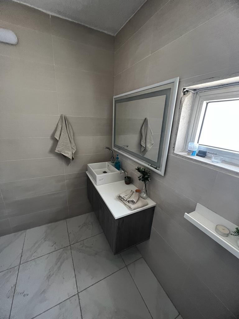 A modern bathroom is presented with light-colored walls and a spacious vanity featuring a sink. A large mirror above the sink reflects the area, while a window allows natural light to brighten the space. Neutral towels hang on the wall, adding to the simplicity.