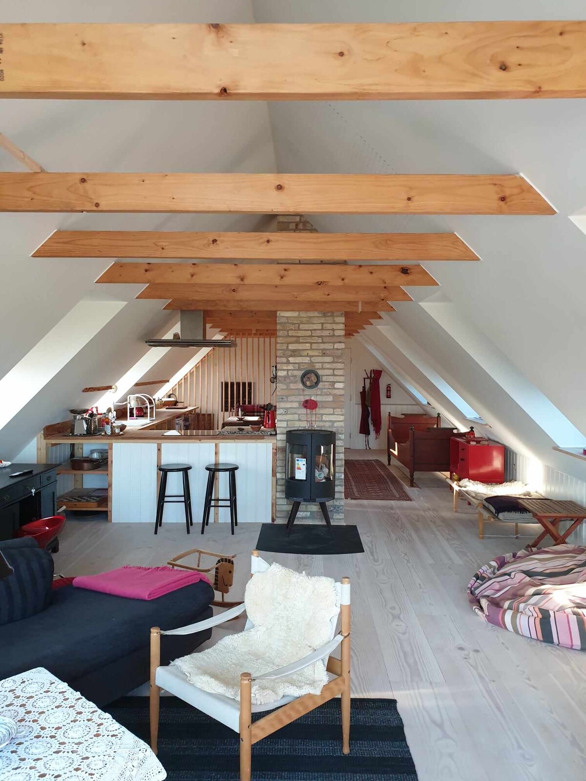 An open living area features a vaulted ceiling with wooden beams, enhancing the spaciousness. A kitchen with modern appliances is visible, alongside a cozy seating arrangement. A brick fireplace adds warmth, while various seating options, including a large cushion and a textured armchair, create a relaxed atmosphere.