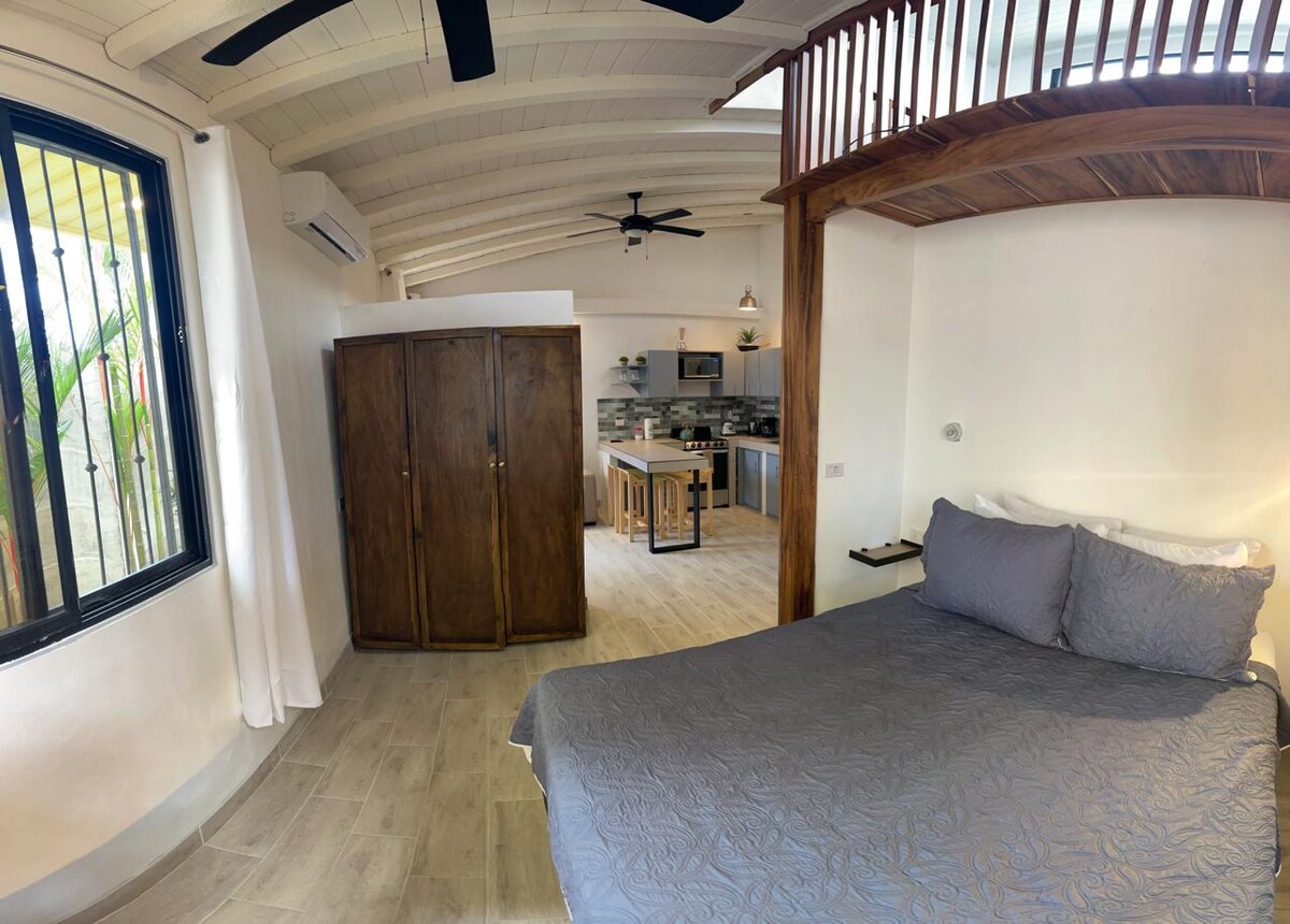 An open concept mini apartment features a spacious sleeping area with a comfortable bed dressed in a light-colored coverlet. A wooden wardrobe is situated nearby, while the kitchen and living area are visible in the background. Large windows allow natural light and views of greenery.