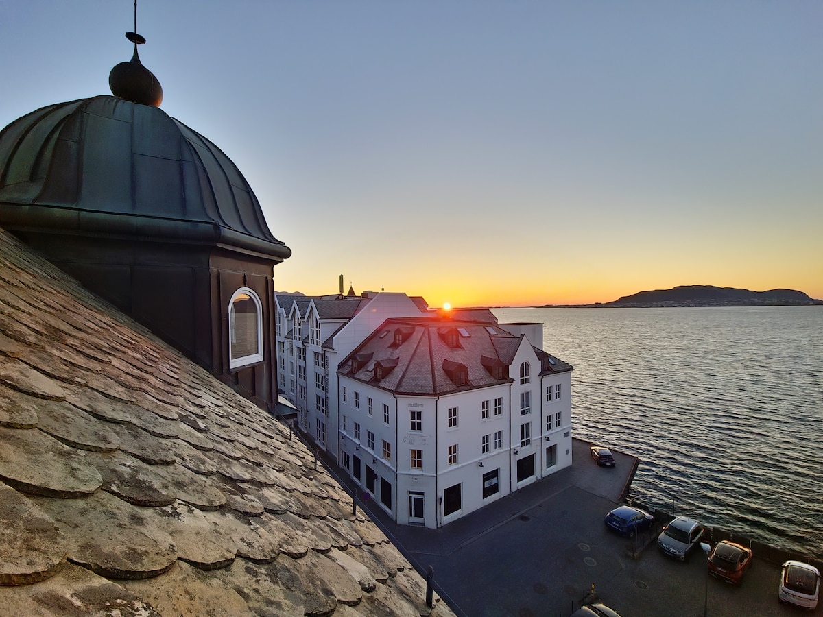 A panoramic view captures the vibrant hues of sunset over the water. The image features the silhouette of a domed rooftop, with a historic building in the foreground and calm waters reflecting the warm tones of the sky.