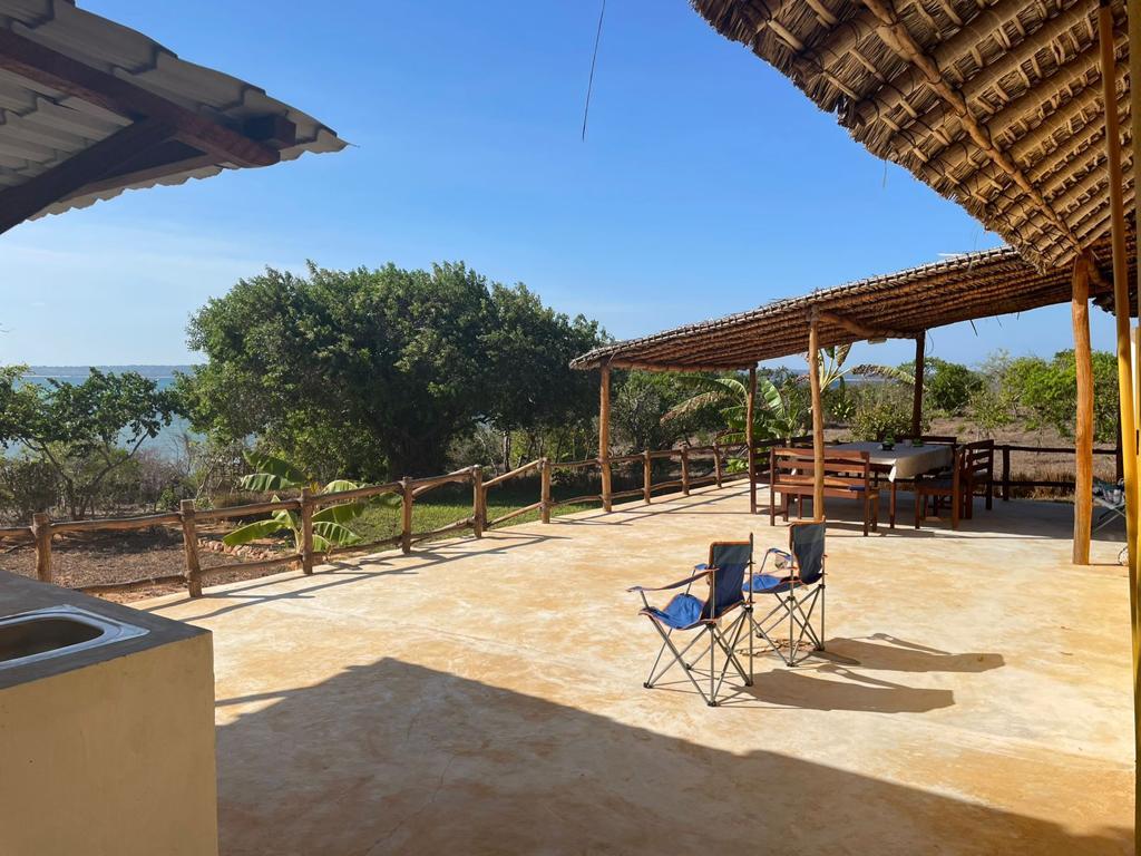 An outdoor seating area features two folding chairs positioned on a spacious patio. A bamboo-covered structure provides shade, while lush greenery borders the scene. The distant view includes a clear sky meeting the ocean, enhancing the serene setting.