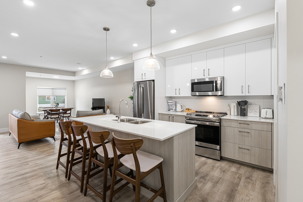 An open kitchen space showcases sleek cabinetry and stainless steel appliances, including a modern refrigerator and oven. A large island with seating for four is highlighted, while natural light filters through a nearby window, illuminating the dining and living area in the background.