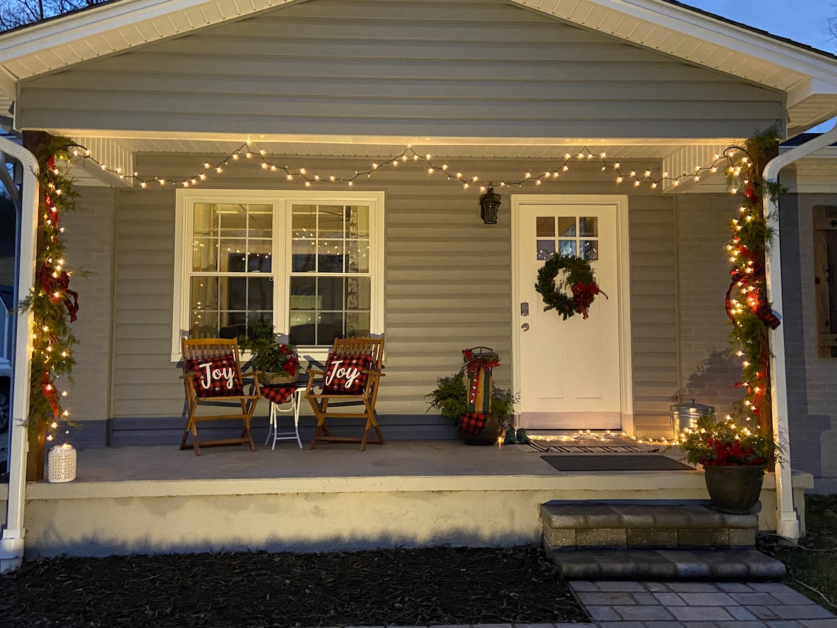 The welcoming front porch features two wooden chairs adorned with festive 'Joy' pillows. String lights illuminate the space, highlighting wreaths on the doors. Potted plants and seasonal decorations enhance the inviting atmosphere, creating a pleasant entryway experience.