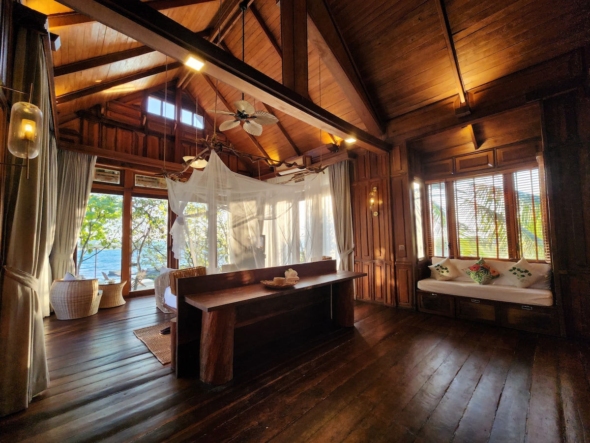 The spacious interior features wooden walls and beams, with natural light filtering through large windows. A seating area is present, along with a bed draped in white linens beneath a mosquito net. Decorative pillows add color to the relaxed seating nook.