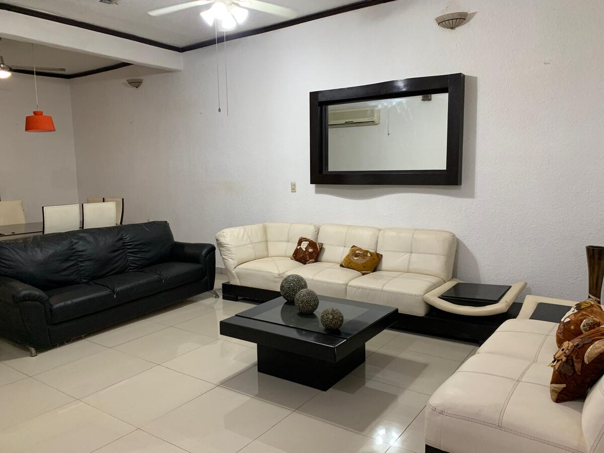 A living area is furnished with a black leather couch and a white sectional sofa, both arranged around a black coffee table. Decorative items are placed on the table, and a large mirror enhances the space's openness. Soft lighting complements the neutral wall tones.