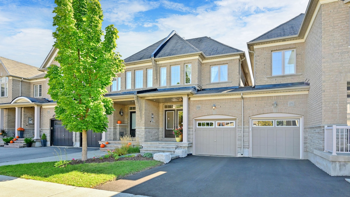 A spacious townhouse exterior is showcased, featuring a well-maintained façade with large windows. A manicured lawn and a young tree contribute to the curb appeal. The driveway is smooth and leads to two garage doors, providing convenience for parking.