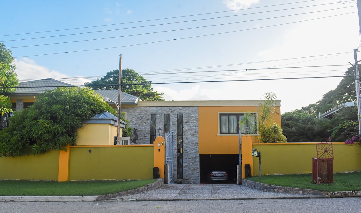 A modern, two-story home features a striking yellow and stone façade. A driveway leads to a covered garage underneath the main structure. Lush greenery surrounds the property, enhancing the inviting appearance of the entrance gate, which opens to a well-kept yard.