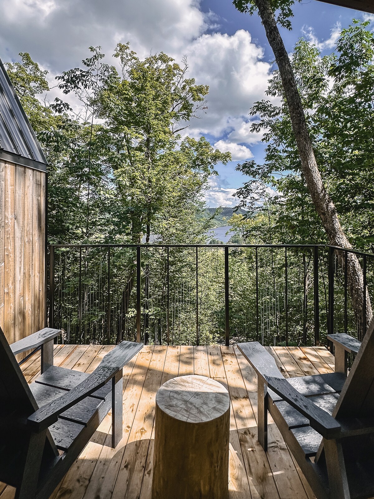 A small covered terrace is surrounded by greenery, featuring two Adirondack chairs positioned around a wooden stump table. The view beyond includes trees and a glimpse of the Saint-Maurice River under a partly cloudy sky.
