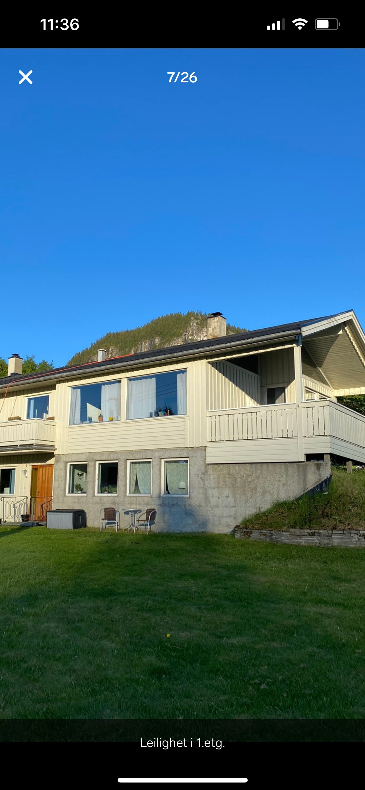 A two-story residence is set against a backdrop of green hills. Large windows offer views of the outdoors, while a small patio area with chairs is visible on the ground level. The exterior features a mix of wood and white paneling, enhancing the home's natural appeal.