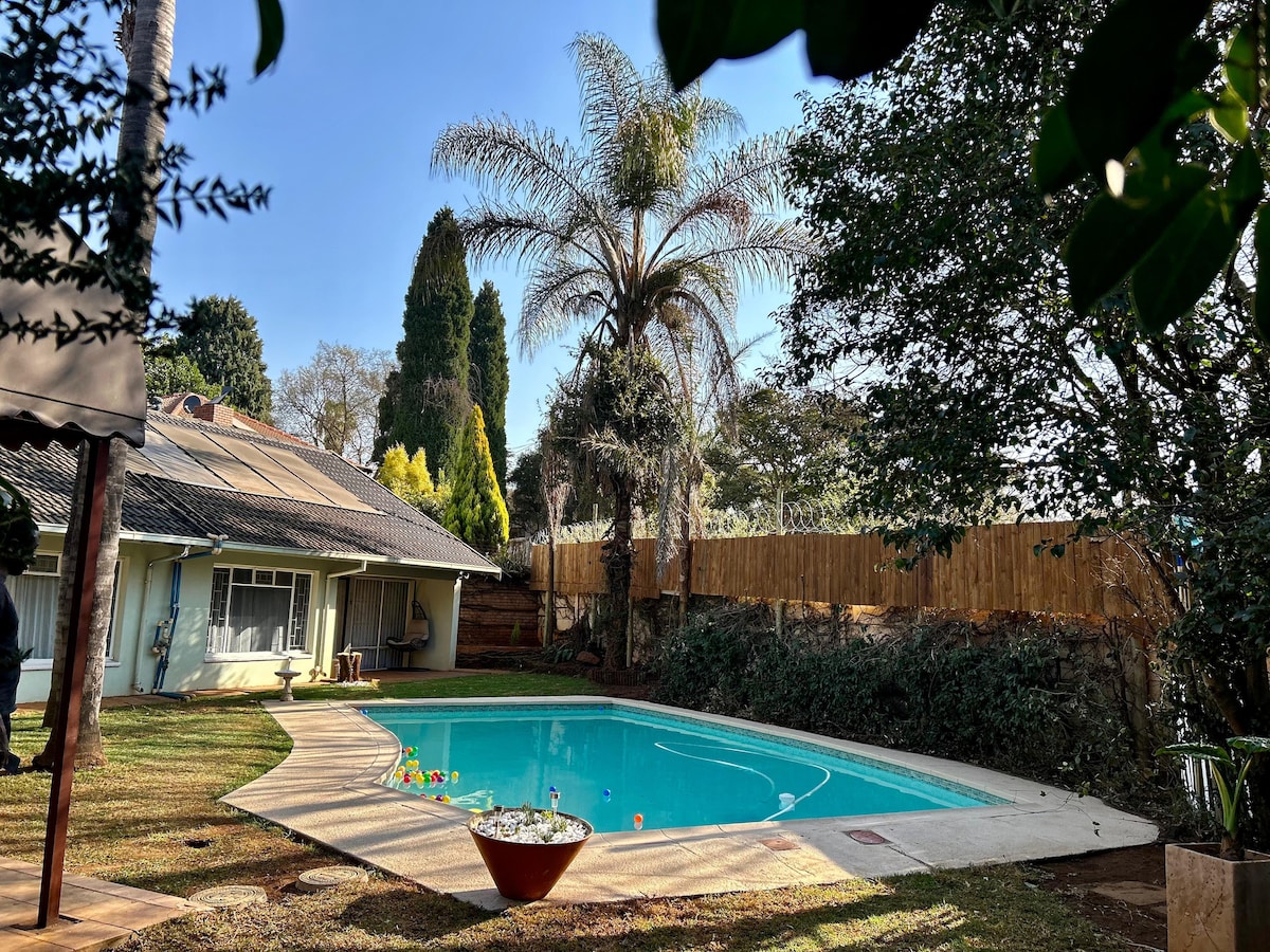A spacious pool area is framed by lush greenery and tall palm trees, providing a private atmosphere for relaxation. The pool, with a clear blue surface, is surrounded by a neatly maintained lawn and features decorative elements along the edge.