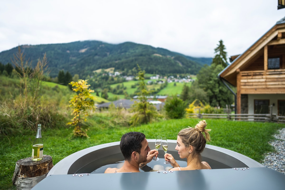 A spacious outdoor hot tub is featured on the terrace, offering scenic views of the lush green mountains and valleys. Two glasses of champagne rest in the hands of two individuals, enhancing a feeling of relaxation and tranquility in this natural setting.