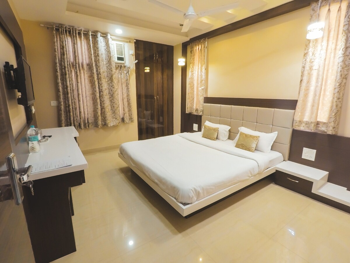 A modern bedroom features a spacious bed adorned with white linens and accent pillows. Two curtains frame the windows, allowing soft light to enter. A minimalist desk and entertainment unit are positioned against the wall, while warm lighting enhances the calming atmosphere.