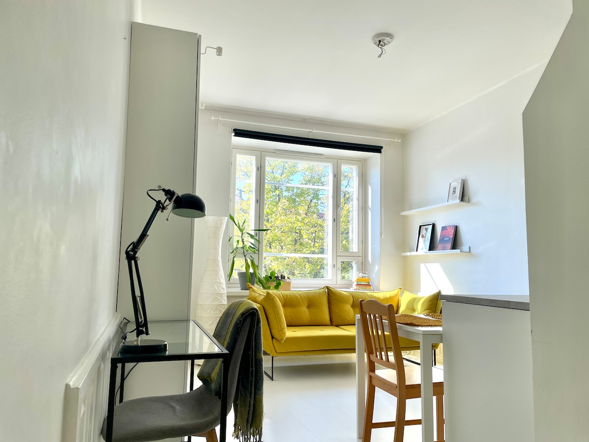 A well-lit living space features a large window allowing ample natural light. A cheerful yellow sofa is situated alongside a wooden dining table. A desk with a lamp and a tall plant adds comfort to the area. Simple shelves display neatly arranged items.