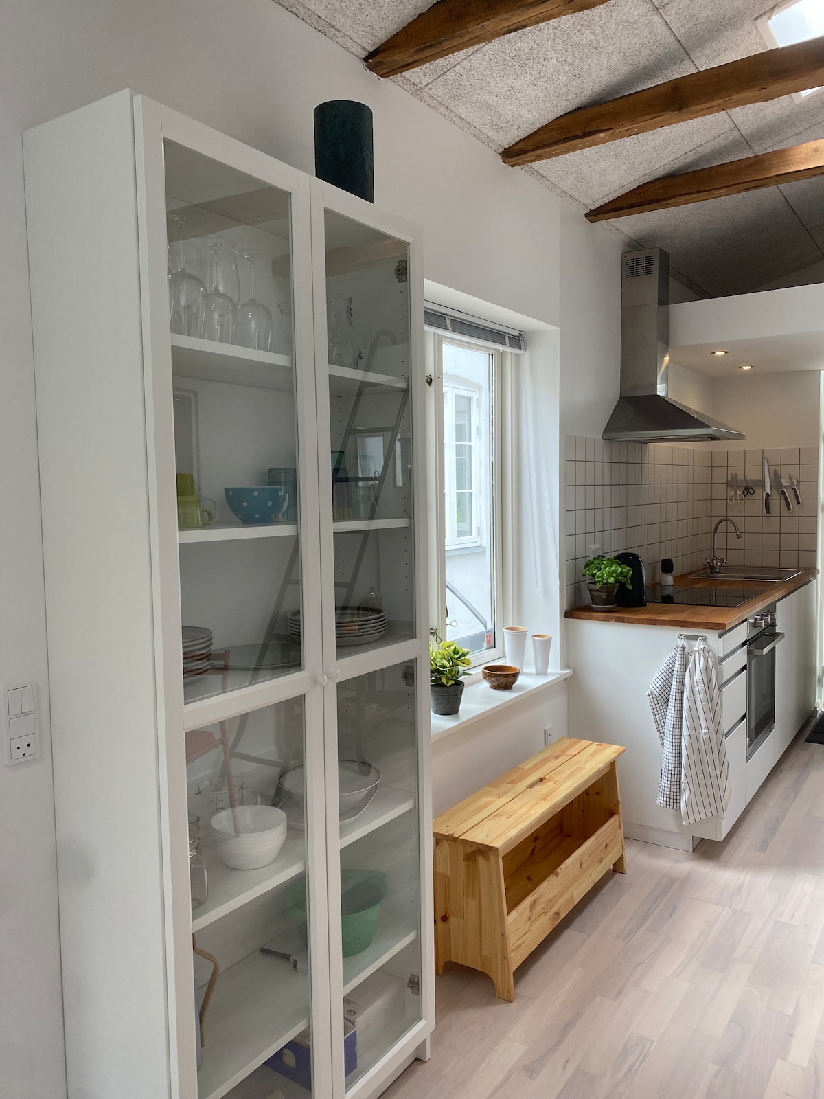 A modern kitchen showcases a glass-fronted cabinet filled with various dishware and glassware. The space features clean white walls and a wooden countertop with a stove and modern appliances. Natural light filters through a nearby window, enhancing the inviting atmosphere.
