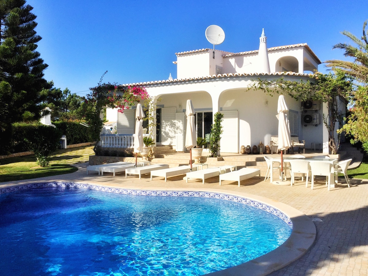 A well-maintained villa features an inviting private pool surrounded by sun loungers and shaded areas. The exterior showcases a spacious terrace with outdoor dining spaces. Lush greenery and mature trees enhance the garden, while the light-colored façade of the house reflects the sunny atmosphere.