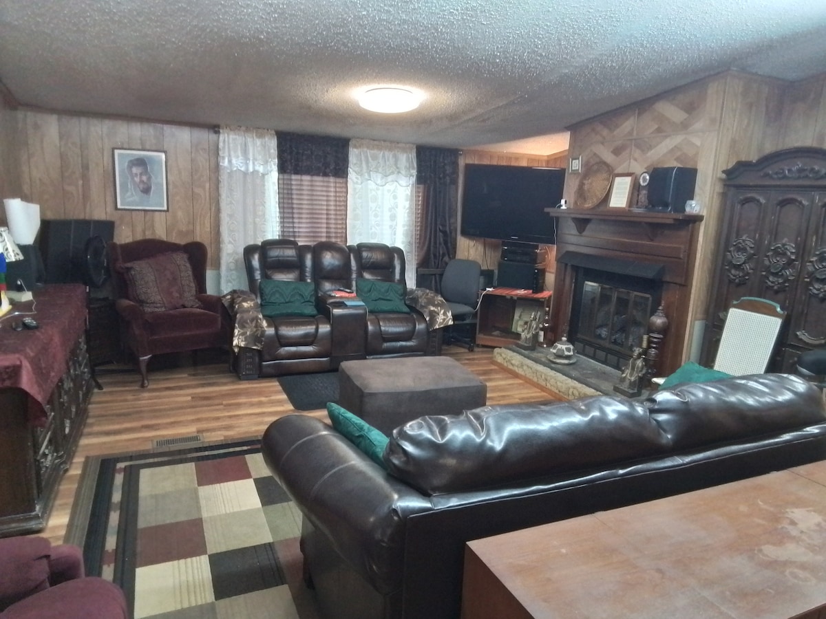 A cozy living room features a mix of comfortable seating, including leather sofas and armchairs. A large coffee table sits centrally on a patterned rug. A fireplace is framed by decorative woodwork, with light filtering through sheer curtains at the windows.
