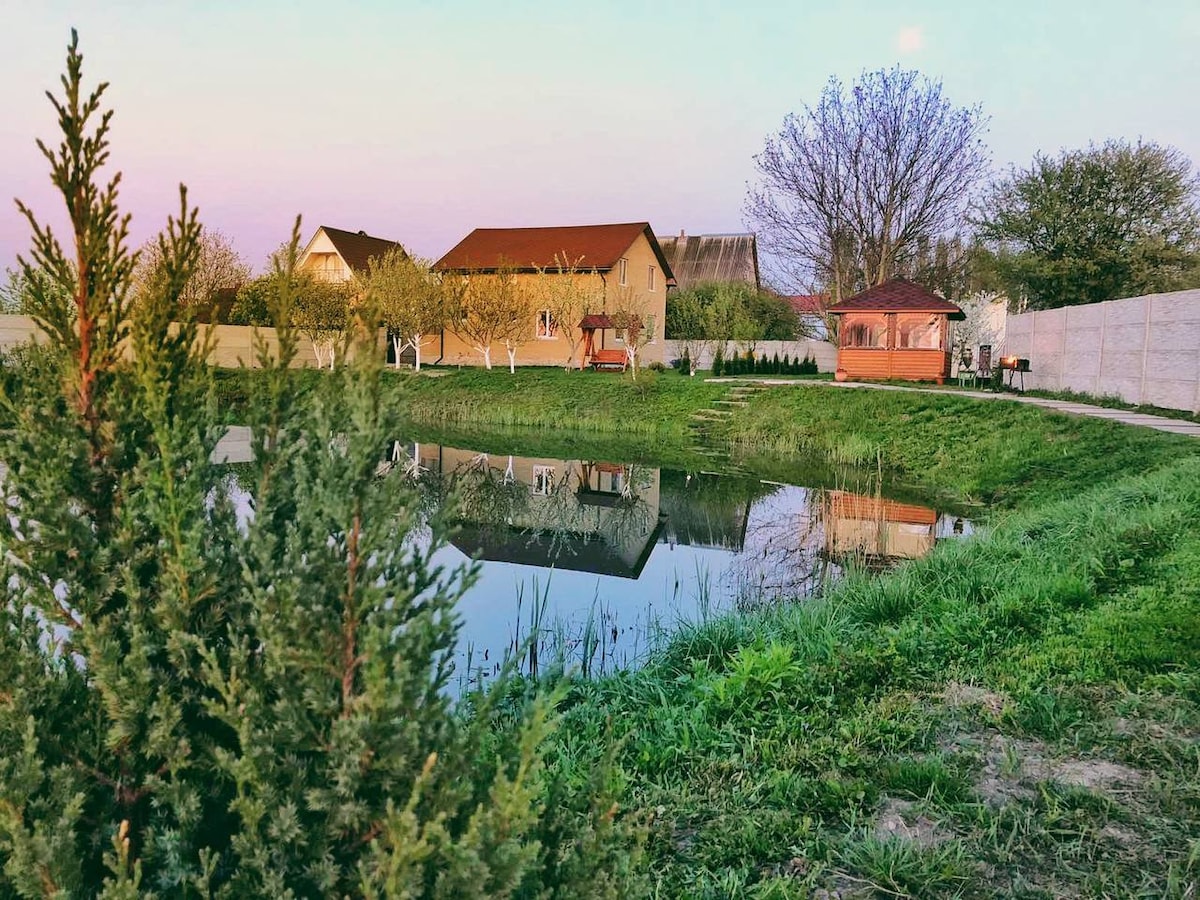 A serene landscape featuring a small pond reflecting the surrounding structures and trees. Green grass and a few scattered trees frame the scene, with a gazebo positioned nearby. The area is ideal for relaxation, surrounded by natural beauty.