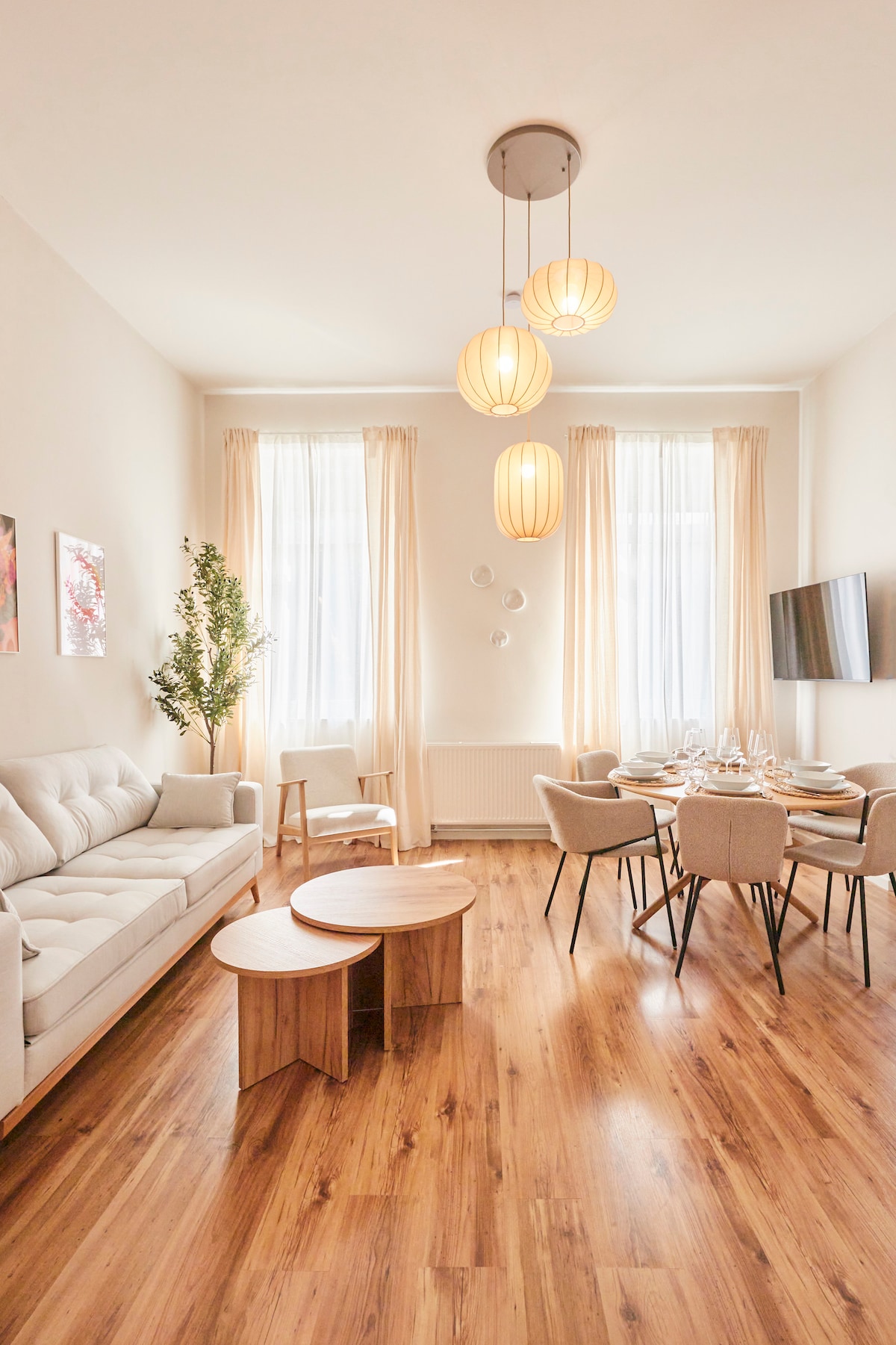 A bright living space is highlighted by natural light streaming through large windows, complemented by soft curtains. A comfortable sofa faces a modern dining table, surrounded by chic chairs. Two wooden coffee tables add a warm touch to the wooden floor, while pendant lights provide subtle illumination.