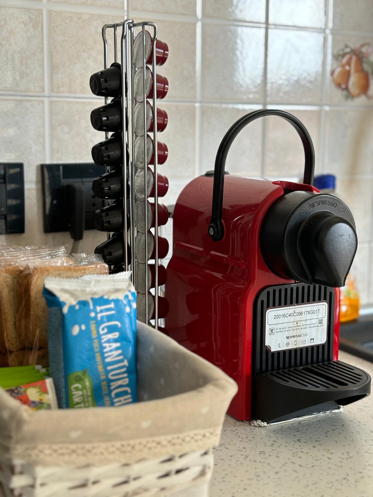A red coffee machine is positioned on a kitchen countertop, with a rotating rack of coffee capsules beside it. A woven basket holds packets, showcasing a tidy and functional coffee station. The backsplash features light tiles with subtle patterns.