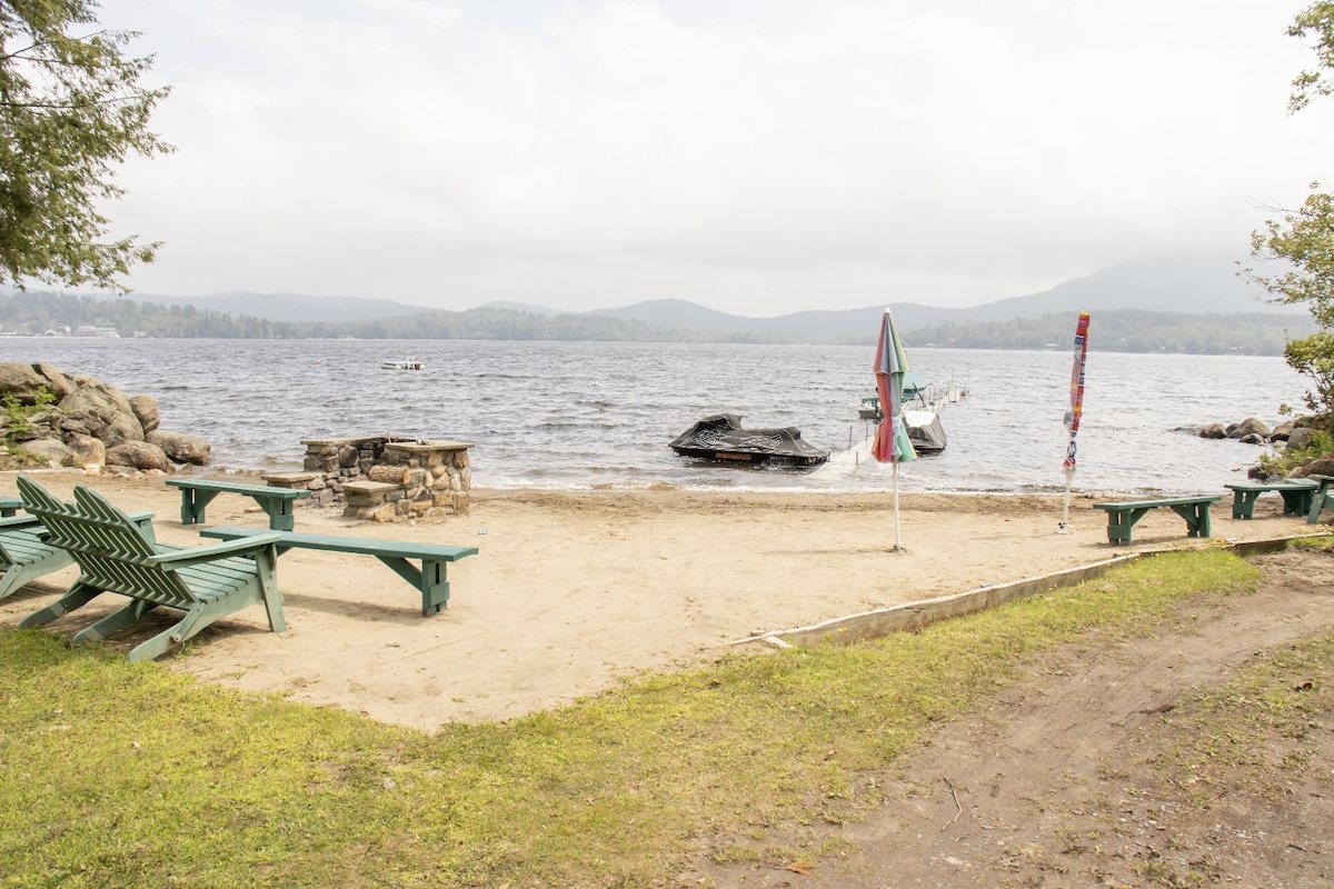 A sandy beach area by Lake Pleasant is surrounded by green Adirondack chairs. A few boats are visible on the water, with rolling hills and a cloudy sky in the background. The tranquil scene offers a relaxing environment for outdoor activities.