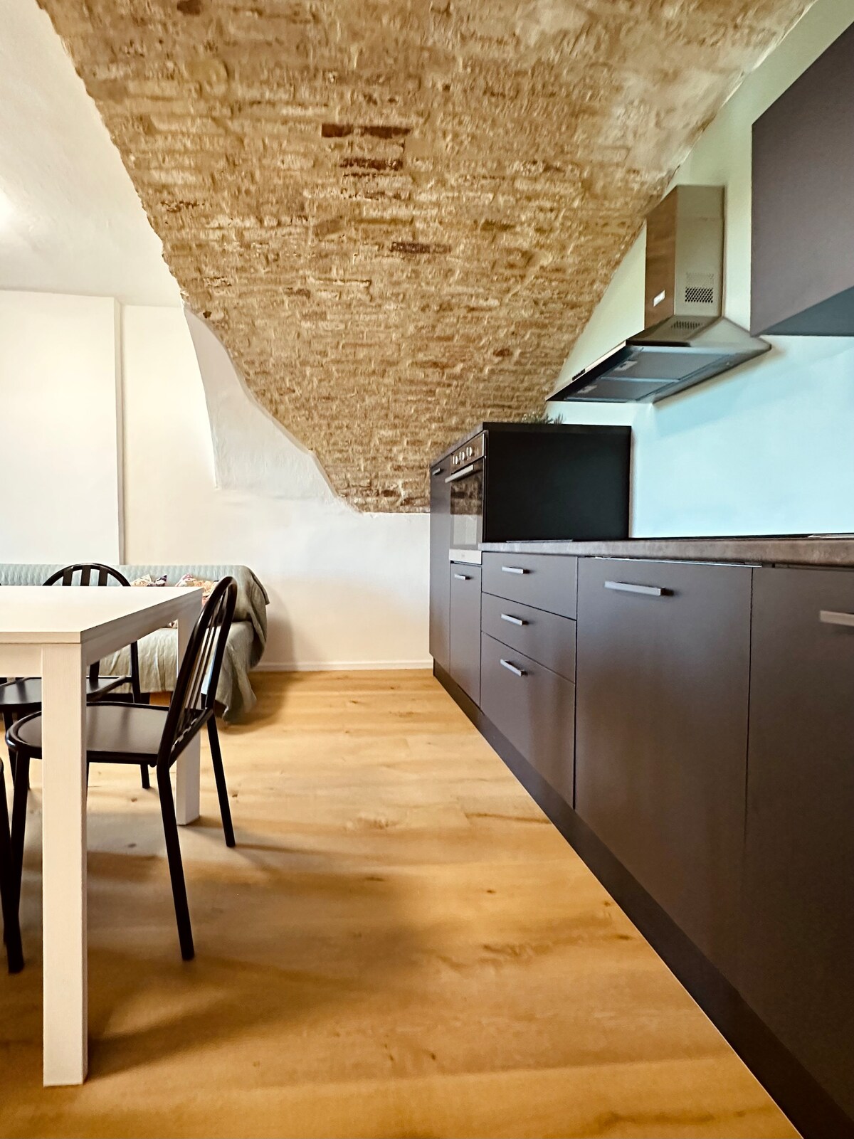 A modern kitchen area features sleek, dark cabinetry and a contrasting light countertop. A rustic brick ceiling adds character above. A dining table with black chairs is positioned nearby, enhancing the kitchen's functional layout.