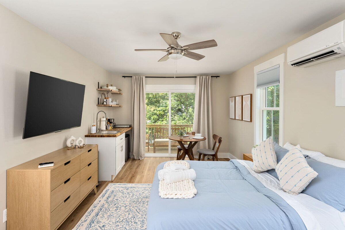A studio space is highlighted with a cozy queen bed dressed in soft linens. A spacious kitchenette is visible, featuring essential appliances. A comfortable dining area for two overlooks the private balcony, while natural light streams through the large windows, enhancing the inviting atmosphere.