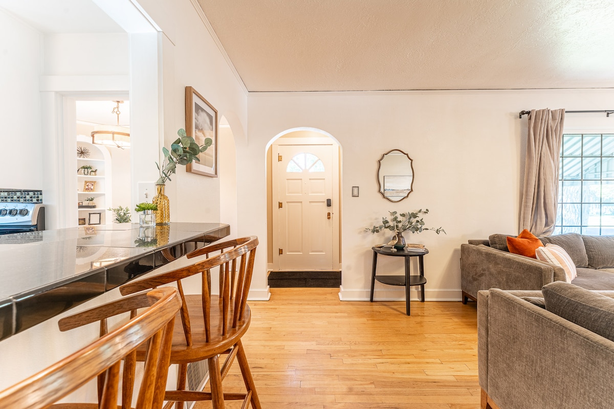 An open living space features hardwood flooring and a cozy seating area with a gray sofa and orange accent pillow. A circular mirror reflects the entryway, and a small table is adorned with greenery. Barstools line the kitchen counter, enhancing the welcoming atmosphere.