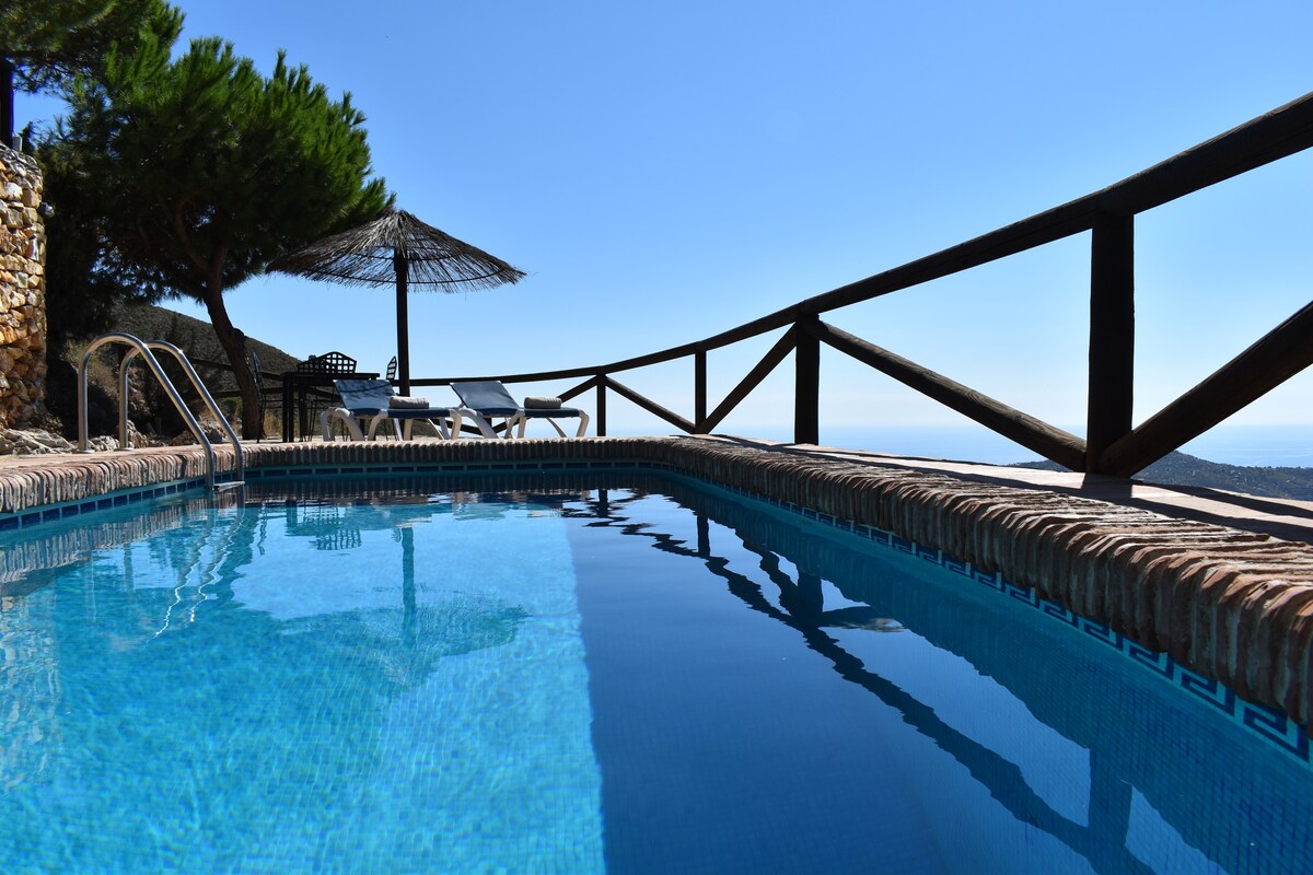 A private swimming pool is surrounded by a natural stone border, reflecting the clear blue sky. A shaded area with a thatched umbrella and sun loungers is visible in the background, offering a tranquil setting with views of the landscape.