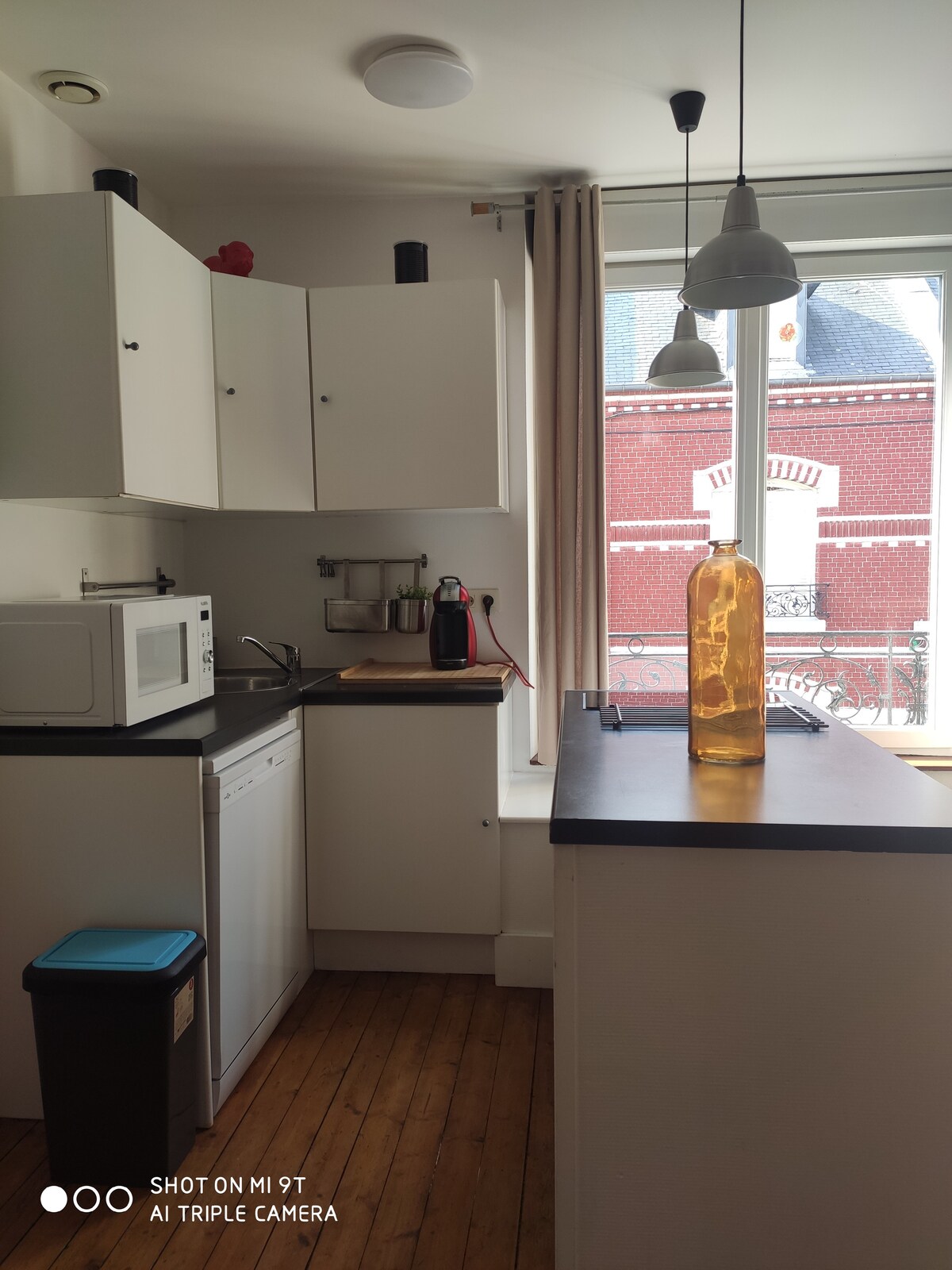 A modern kitchen space is depicted, featuring white cabinetry and a stainless steel dishwasher. A microwave is positioned on the counter near a window, providing natural light. A decorative glass bottle sits on the bar top next to a small stool.