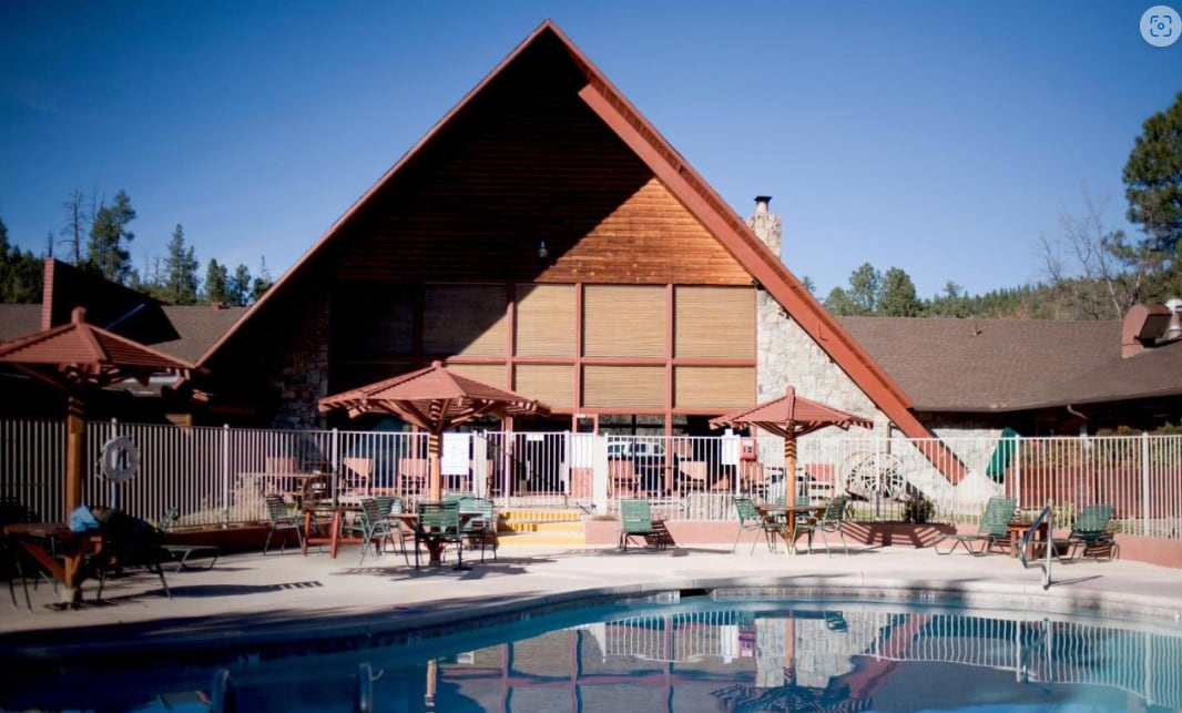 An inviting outdoor pool area features a wooden lodge with triangular architecture in the background. Surrounding the pool, there are multiple shaded seating areas with tables and umbrellas. A wooden fence encloses the space, enhancing the secluded atmosphere.