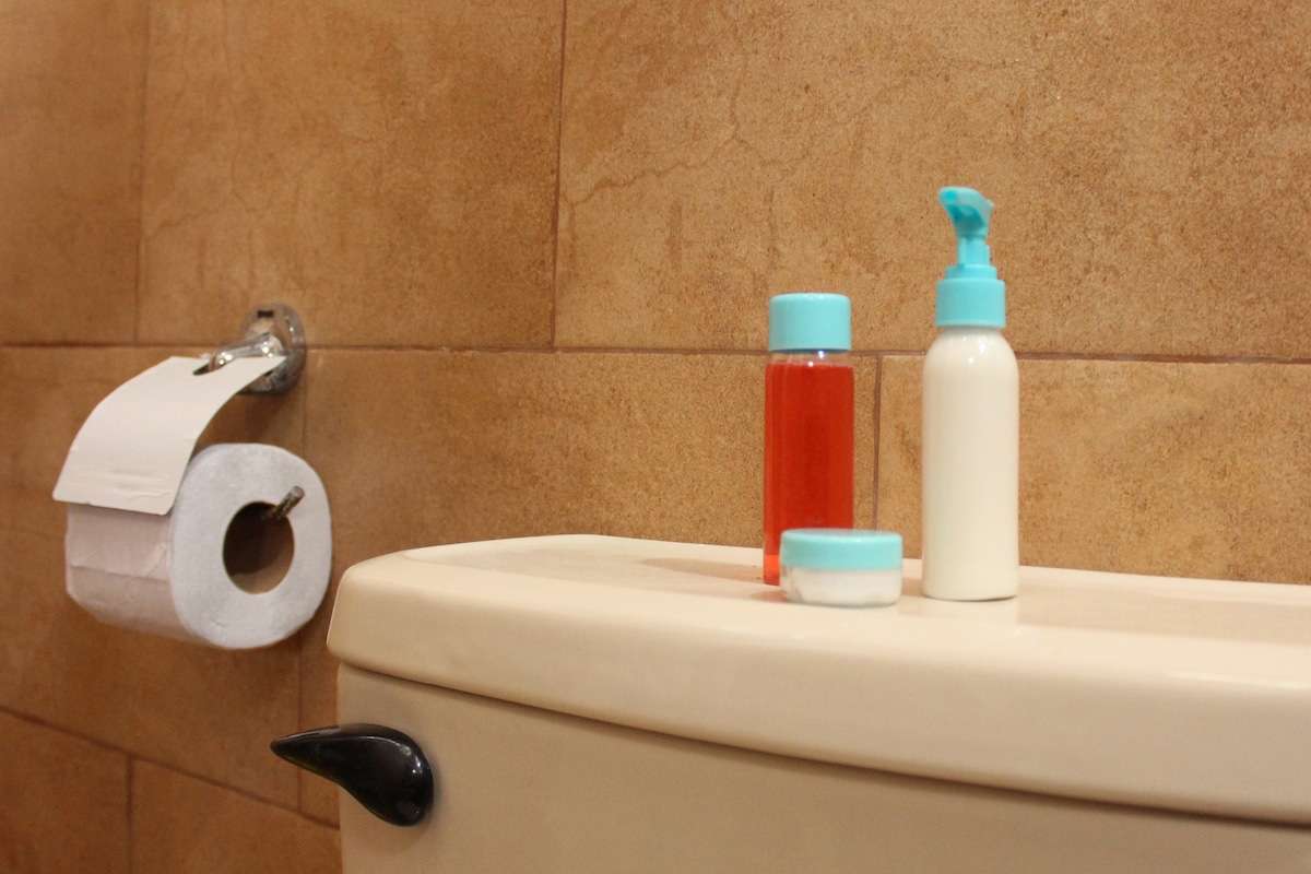 A bathroom setting features a cream-colored toilet tank topped with three personal care items: a small jar, a bottle of lotion, and a shampoo bottle, all with blue caps. A roll of toilet paper is mounted on the wall nearby.