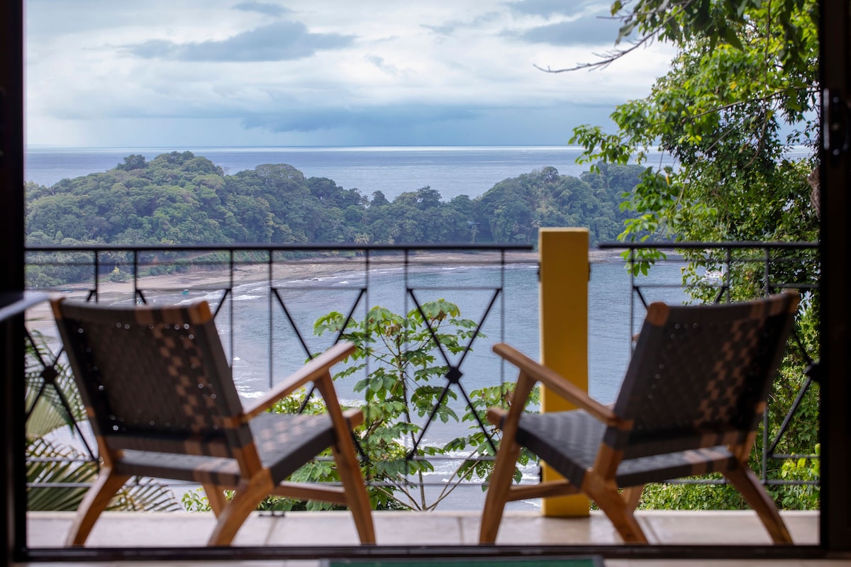 Two wooden chairs are positioned on a balcony, offering a clear view of the ocean and the surrounding lush greenery. The calm waters of the bay reflect the cloudy sky, creating a serene atmosphere. A vibrant yellow post adds a pop of color to the scene.