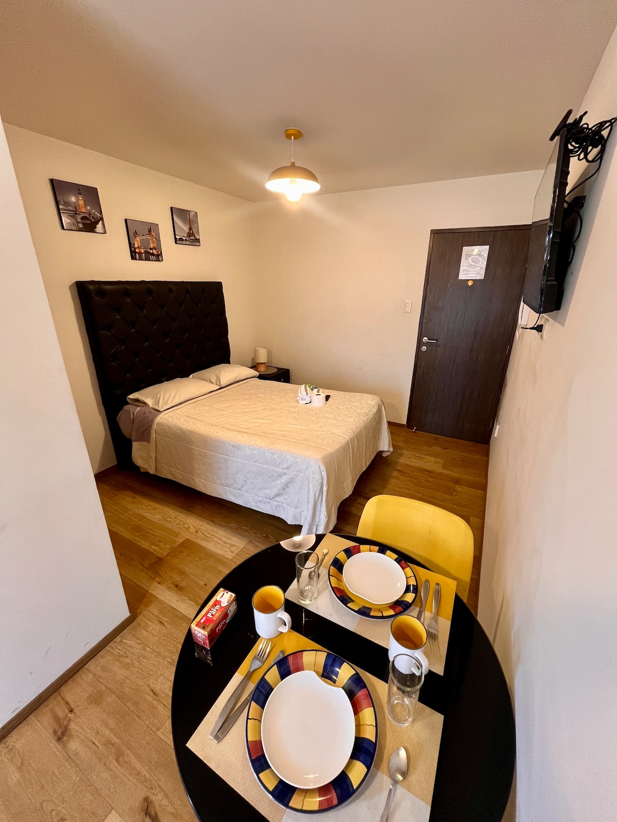A cozy sleeping area features a spacious bed with a light-colored blanket. A small dining table is set for two, displaying plates, utensils, and drinks. The room is illuminated by a ceiling light, while framed artwork adorns the walls.